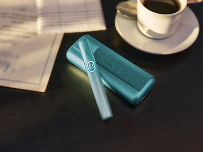 Breeze Blue IQOS ILUMA i Prime holder resting on charger on a workspace with coffee and papers