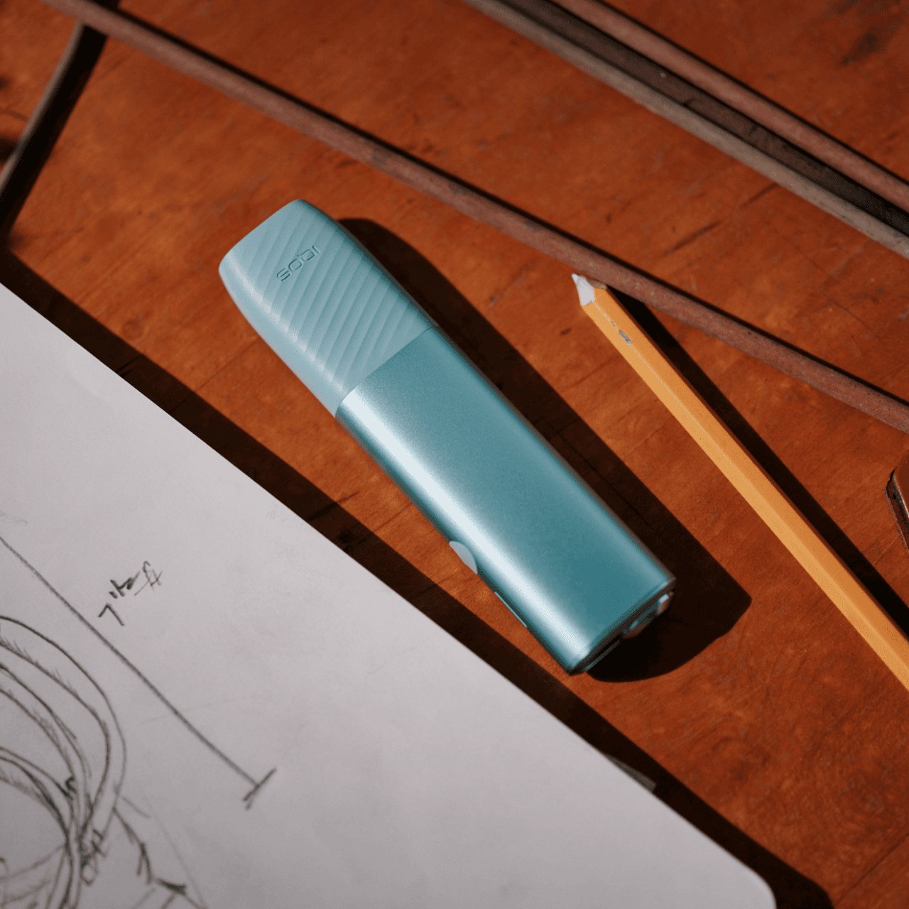 An IQOS ILUMA i ONE color Breeze Blue next to a pencil and a drawing