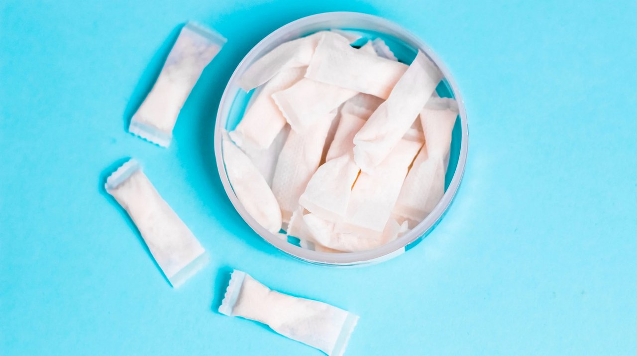 Offene Dose mit weißen Nicotine Pouches auf blauem Hintergrund, einige Pouches liegen neben der Dose.