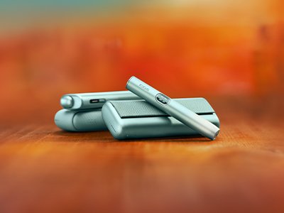Breeze Blue IQOS ILUMA i ONE holder lying on top of a Breeze Blue IQOS ILUMA i charger behind a Breeze Blue IQOS ILUMA i Prime holder resting on charger on a surface against a warm orange background