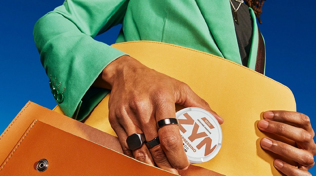 Person steckt eine ZYN-Dose in eine gelbe Ledertasche, vor klarem blauem Himmel. 