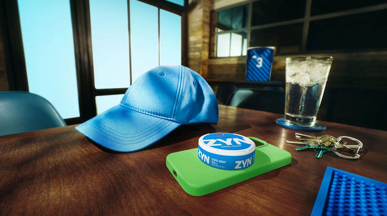 Nahaufnahme eines Holztisches mit blauer Baseballkappe, ZYN-Dose auf grünem Smartphone und Autoschlüssel im Hintergrund