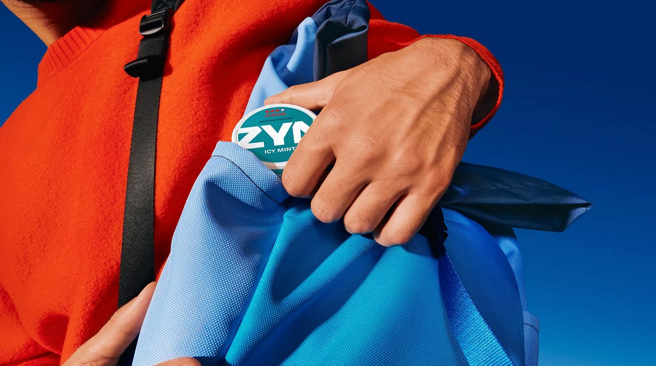 Person steckt eine ZYN-Dose in eine blaue Tasche, vor klarem blauem Himmel, trägt auffälliges rotes Oberteil.