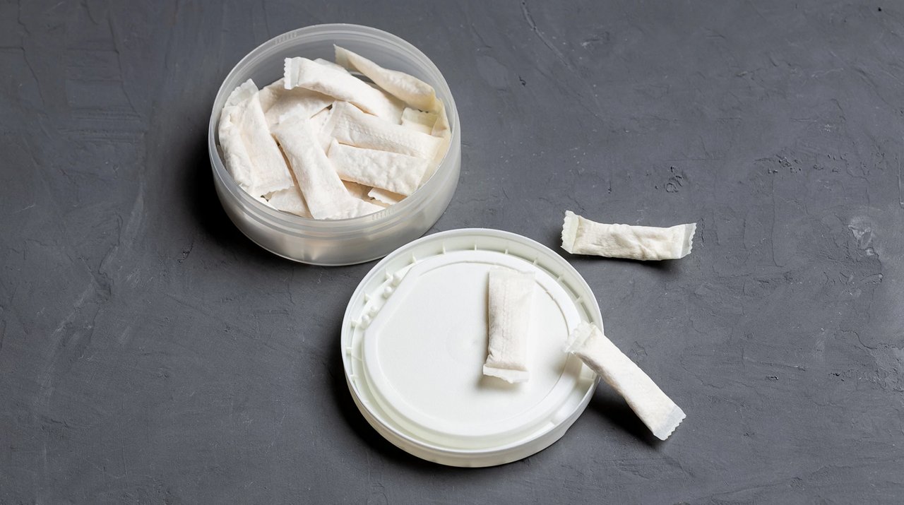 Offene Dose mit weißen Nicotine Pouches auf grauer Oberfläche, Deckel liegt daneben mit Pouch darauf.