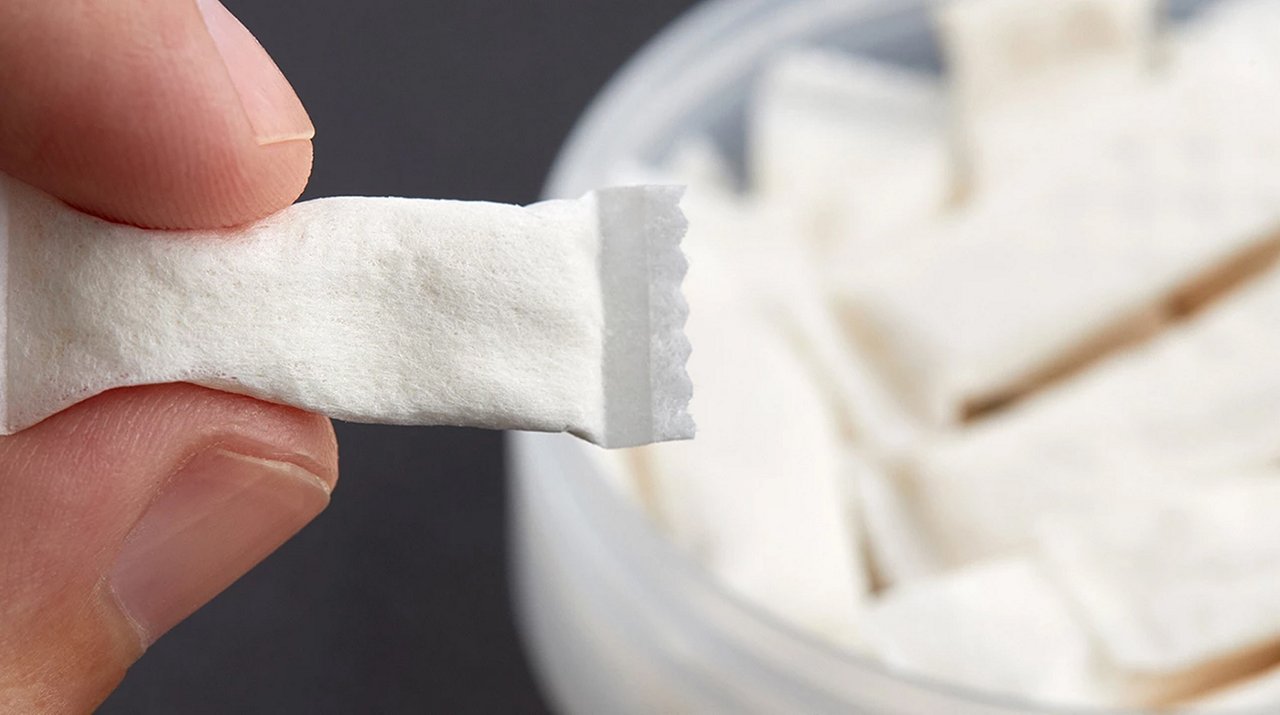 Nahaufnahme einer Hand, die ein Nicotine Pouch hält, im Hintergrund eine geöffnete Dose. 