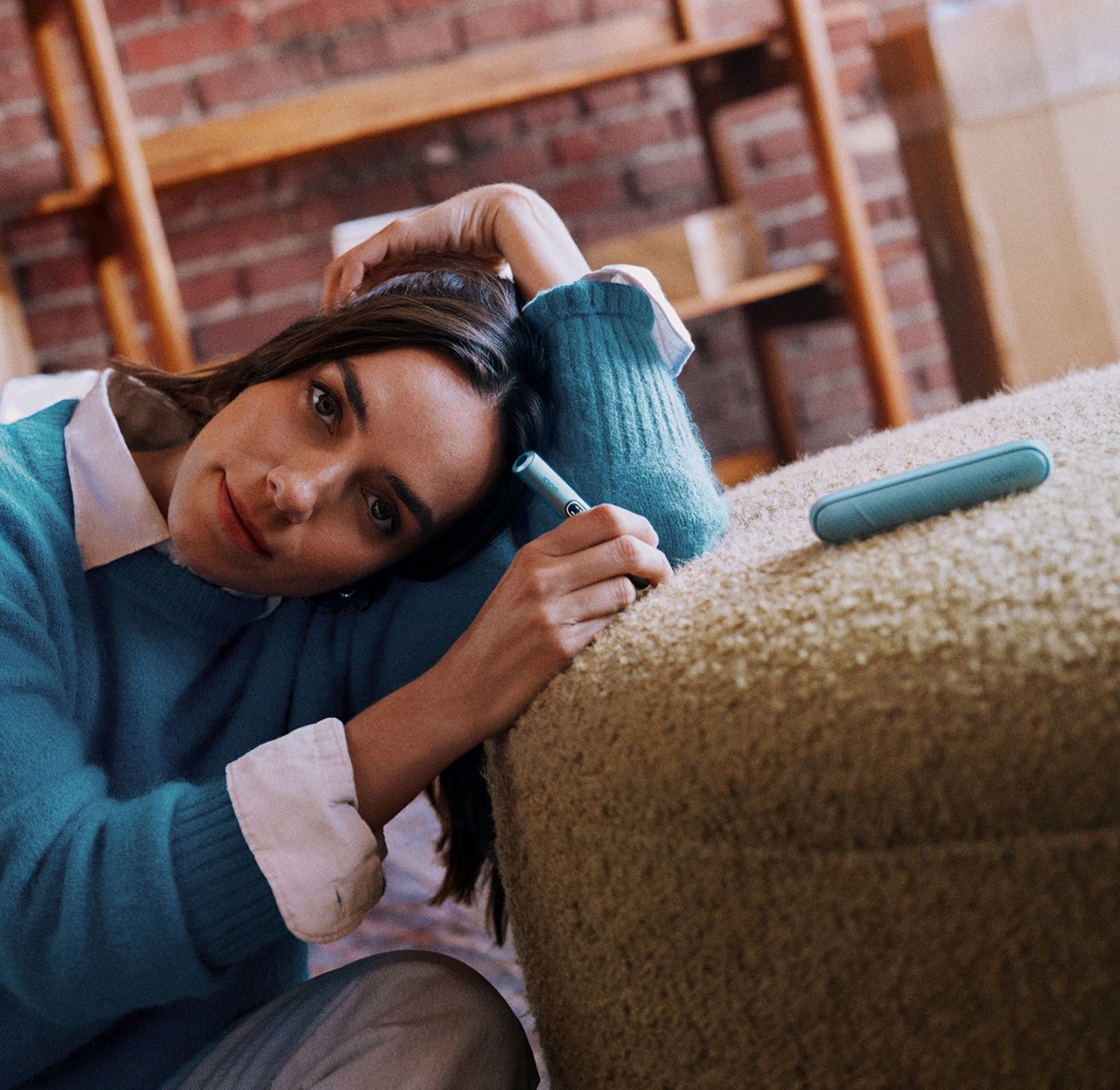 A woman lying on a Sofa and holding an IQOS ILUMA i color Breeze Blue on their hand