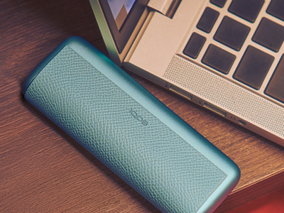 Turquoise IQOS carrying case placed on a wooden desk next to a laptop computer.