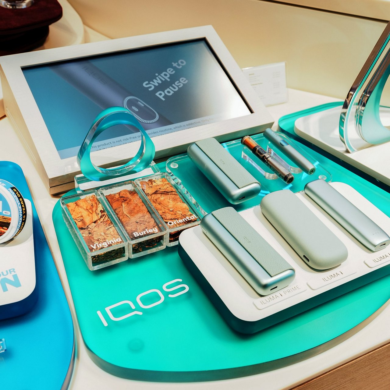 Retail display of IQOS ILUMA i devices with tobacco samples and digital screen and turquoise IQOS key.