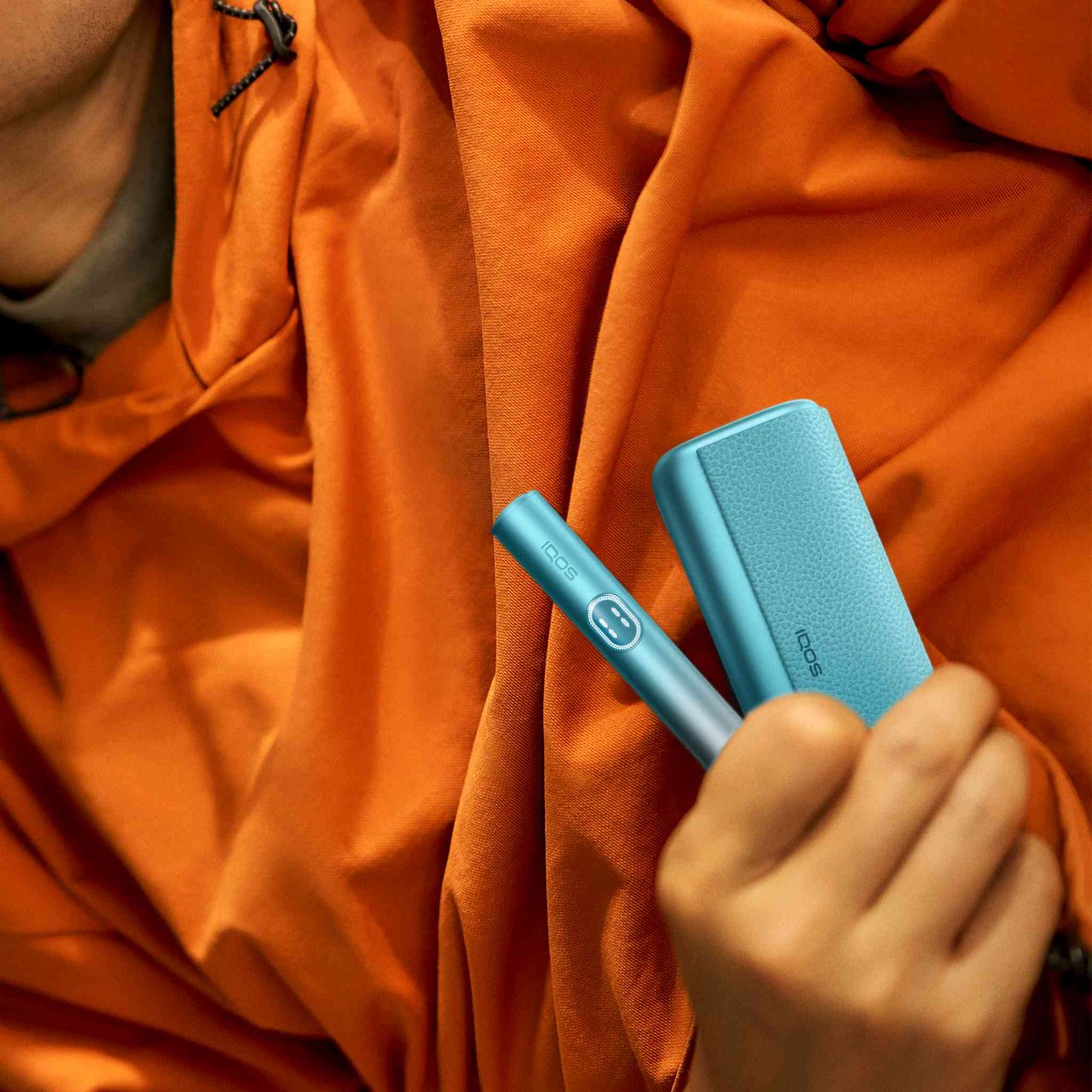 Orange fabric backdrop with visible folds and creases, with a hand holding the new Breeze Blue IQOS ILUMA i PRIME Holder & Pocket Charger.