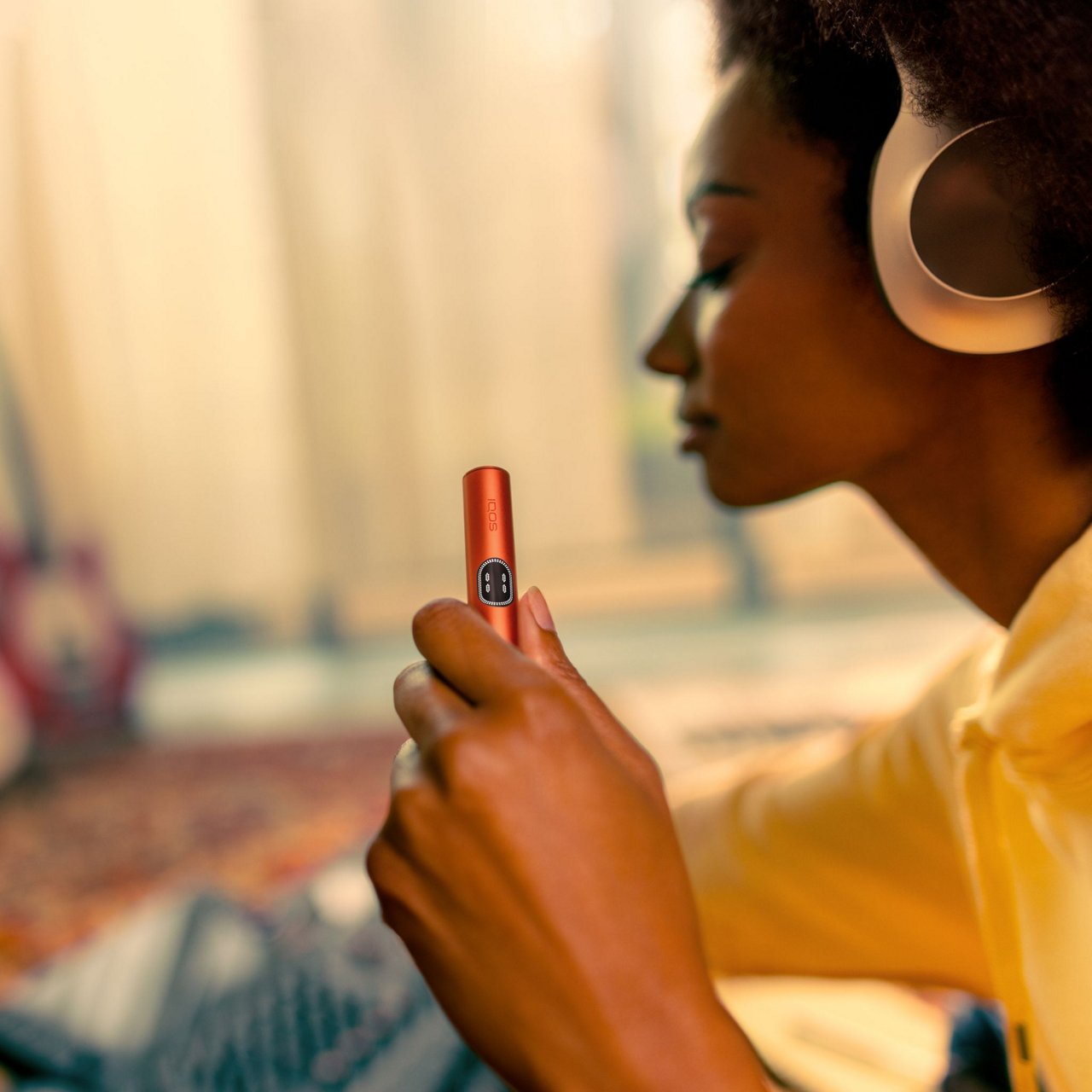 Person wearing headphones holding a red IQOS ILUMA i Holder with a blurred guitar in the background.