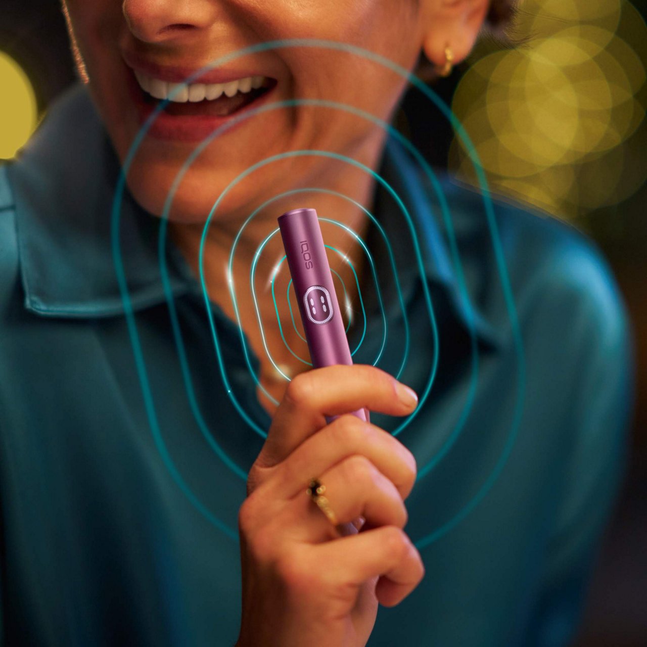 Close-up of a person holding an IQOS ILUMA i PRIME Garnet Red Holder with blue concentric circles indicating vibration, set against a warm, softly blurred background.