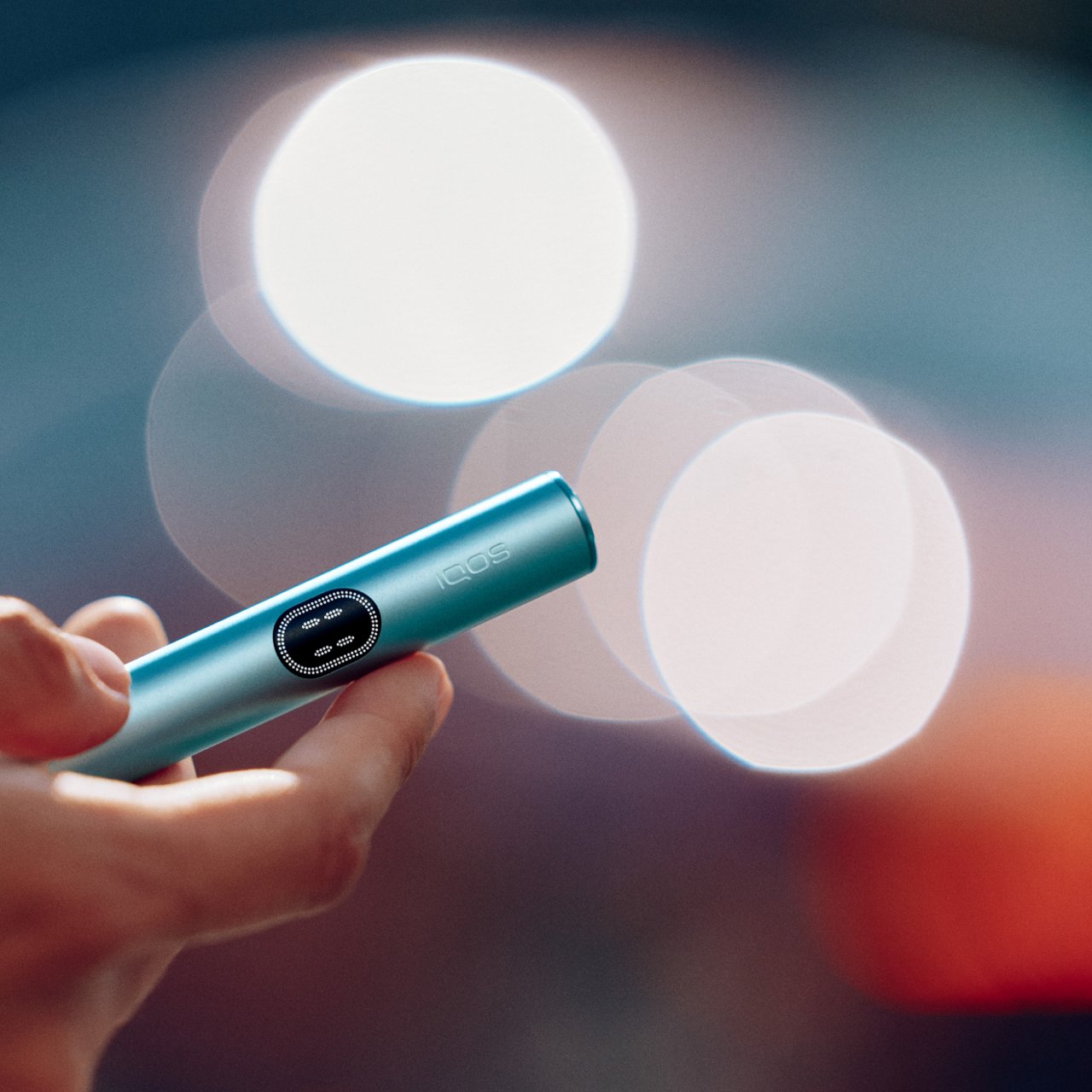 Primer plano de un IQOS ILUMA i One turquesa en la mano con luces desenfocadas al fondo.