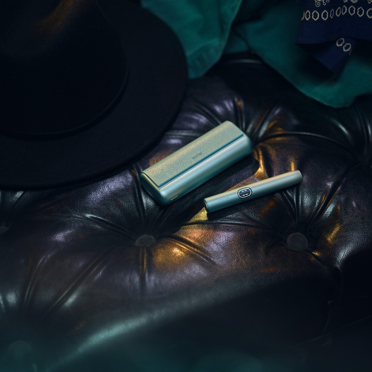 IQOS Iluma i Prime verde colocado sobre sofá de cuero junto a sombrero y prendas