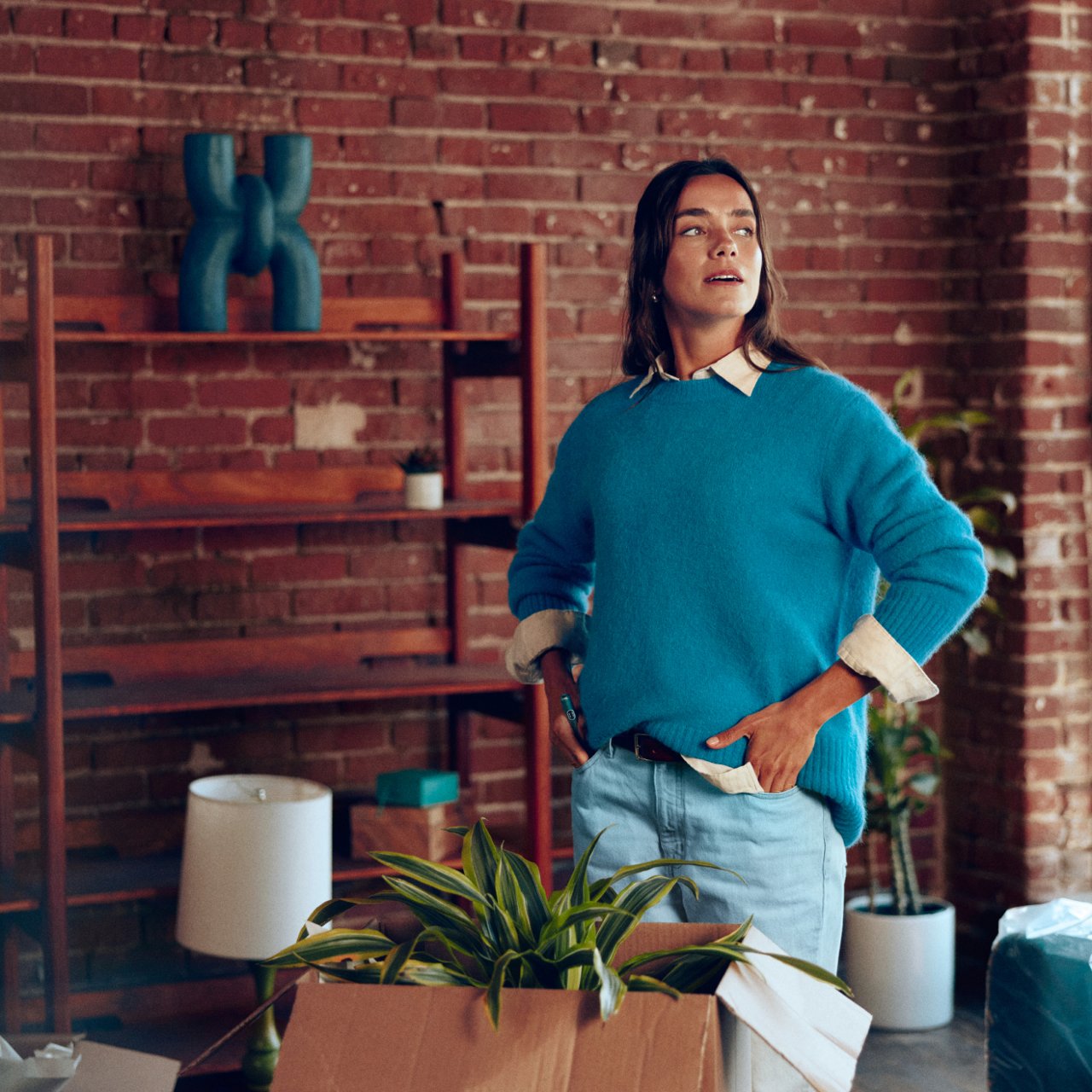 Mujer posando de pie en un interior con plantas y estanterías de fondo, formato grande.