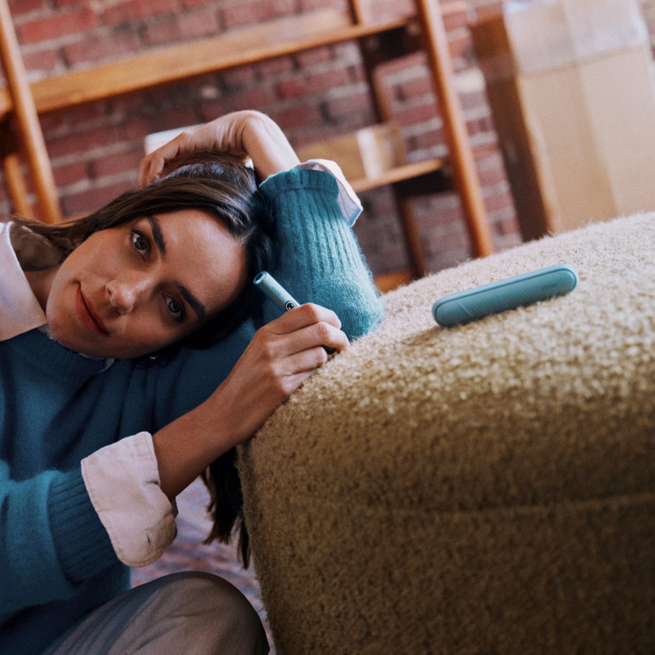 Persona sosteniendo un IQOS ILUMA i One turquesa junto a una laptop sobre una mesa de trabajo.