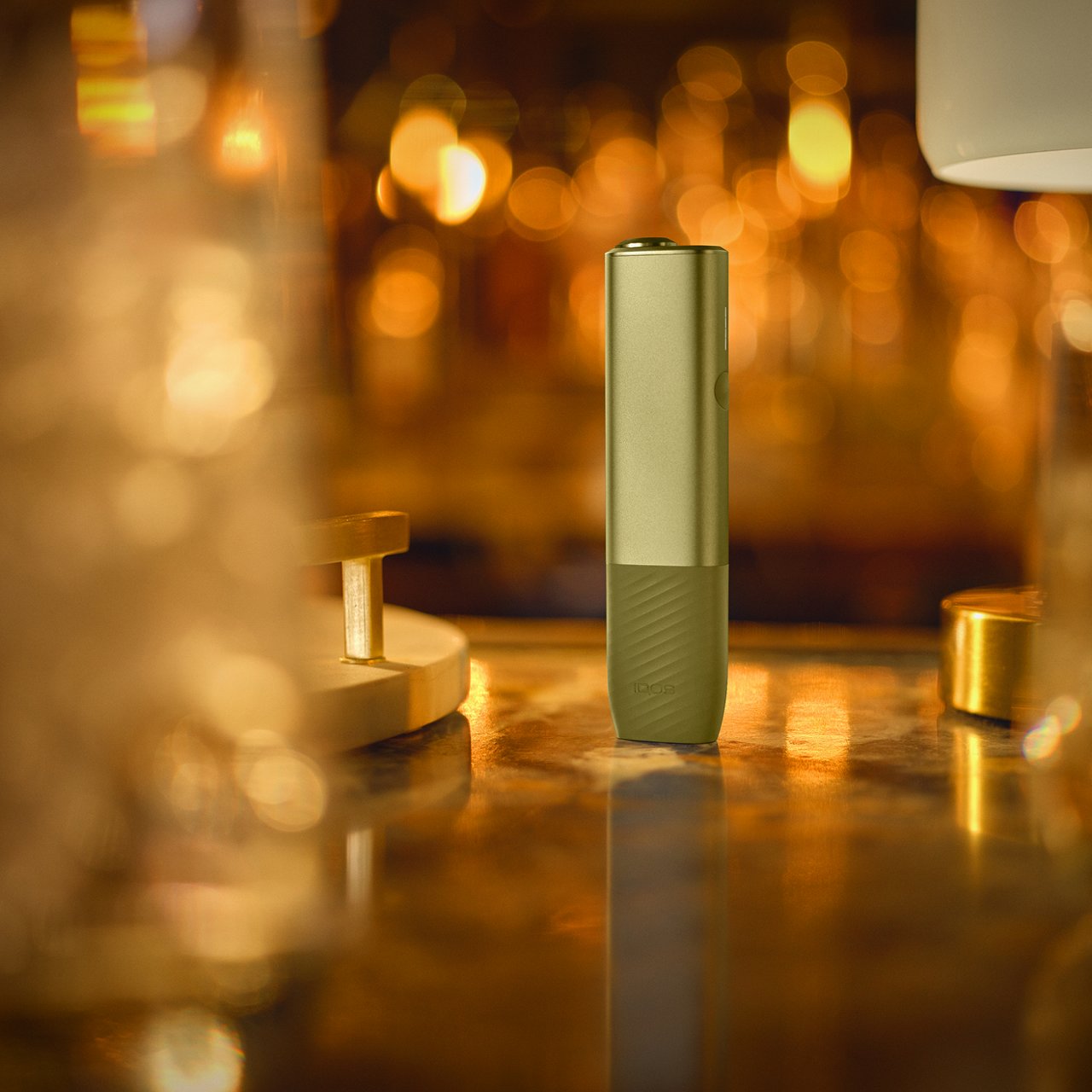 IQOS Iluma i ONE verde en posición vertical sobre mesa iluminada con reflejos dorados