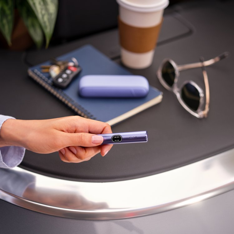 Persona sosteniendo un IQOS ILUMA i One turquesa junto a una laptop sobre una mesa de trabajo.