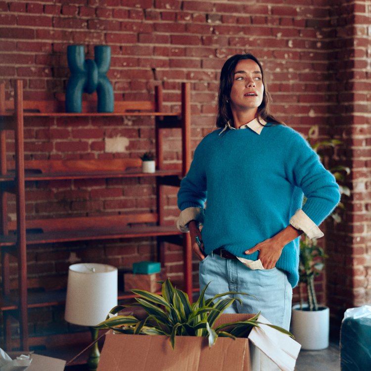 Mujer posando de pie en un interior con plantas y estanterías de fondo, formato grande.