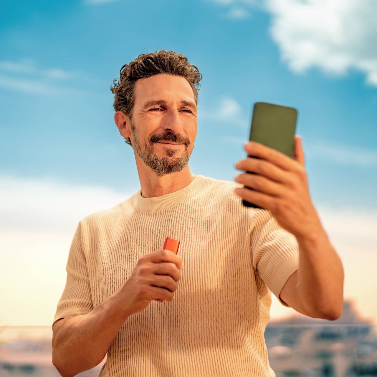 Person holding a smartphone and an IQOS ILUMA i One device outdoors under a clear blue sky with clouds.