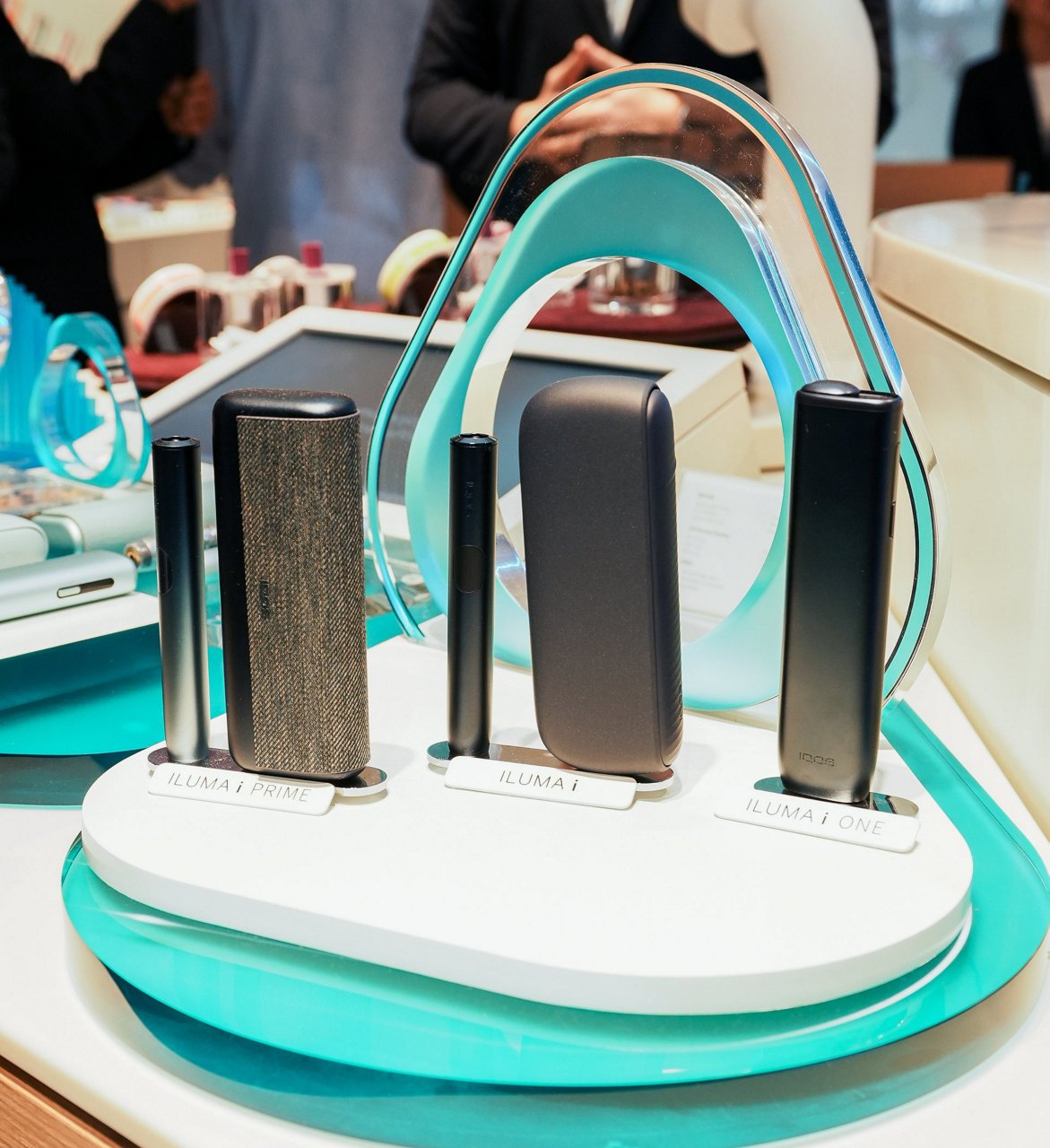 Display of IQOS ILUMA i devices including ILUMA i PRIME, ILUMA i, and ILUMA i ONE showcased on a modern stand with turquoise accents.