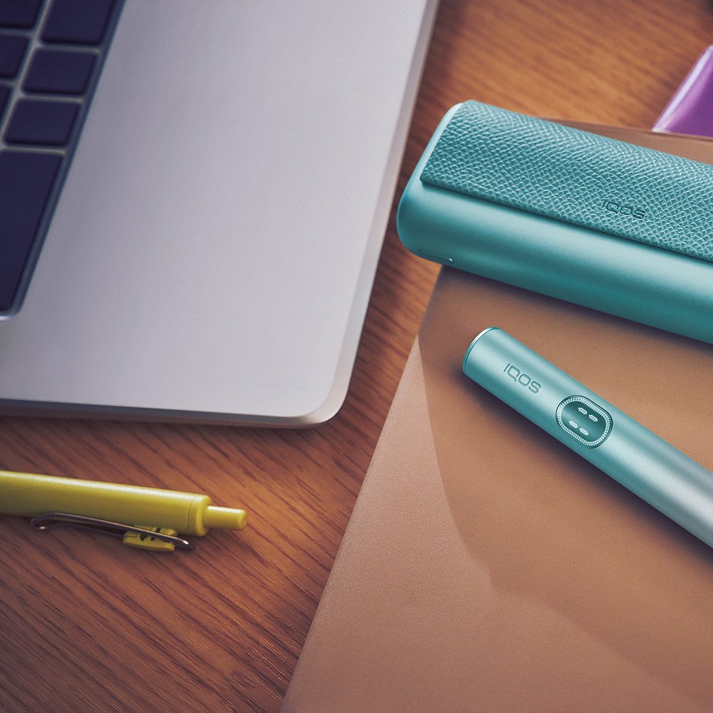 IQOS Tabakerhitzer liegt neben Kit und Laptop auf einem Schreibtisch.