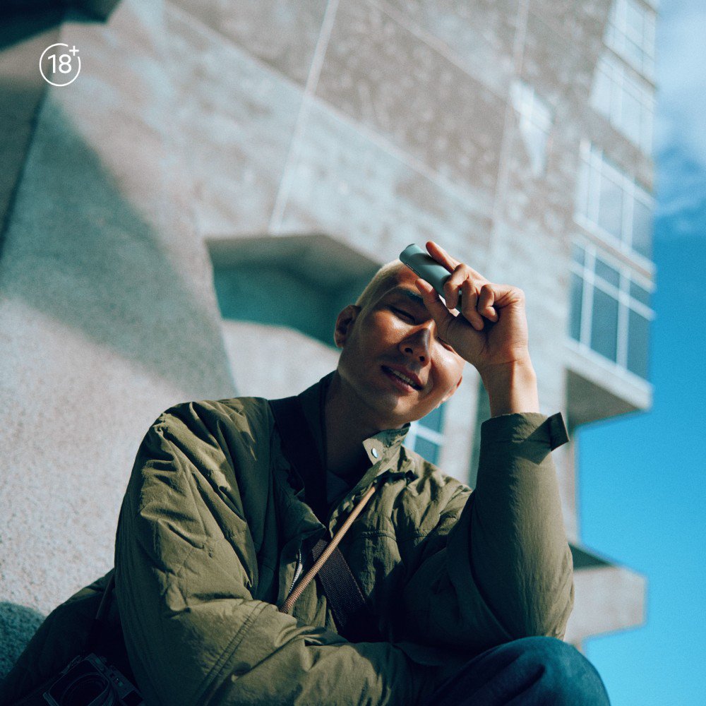 Adult person wearing a green jacket holding an IQOS device outdoors near a modern concrete building under clear blue sky.