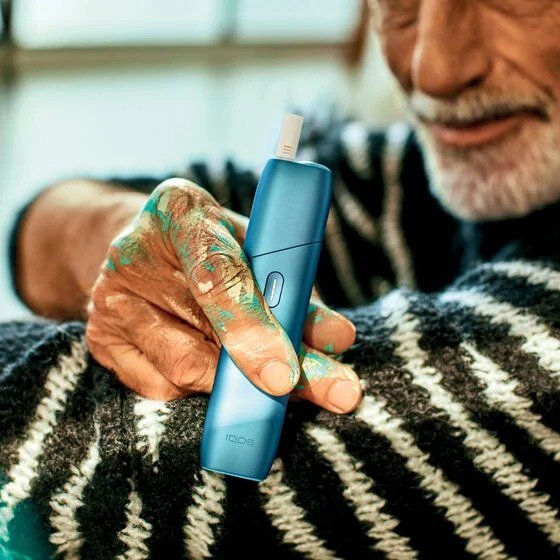 Man with paint on hands holding a turquoise IQOS Originals One heated tobacco device.