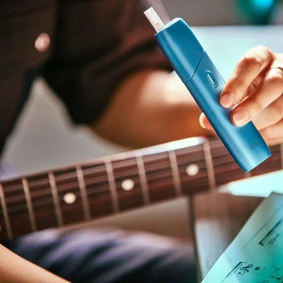 Hand holding a turquoise IQOS Originals One heated tobacco device with a guitar.