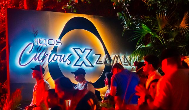 Personas reunidas frente a un letrero iluminado que muestra las palabras ‘IQOS Curious X Zamna’, rodeado de vegetación y luces de colores