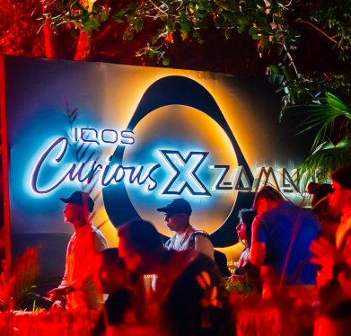 Personas reunidas frente a un letrero iluminado que muestra las palabras ‘IQOS Curious X Zamna’, rodeado de vegetación y luces de colores