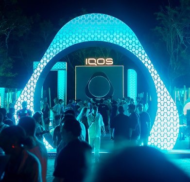 Estructuras iluminadas en tonos azul turquesa formando arcos y columnas, con un letrero de 'IQOS' al fondo y múltiples personas caminando y reunidas en el área durante un evento nocturno.