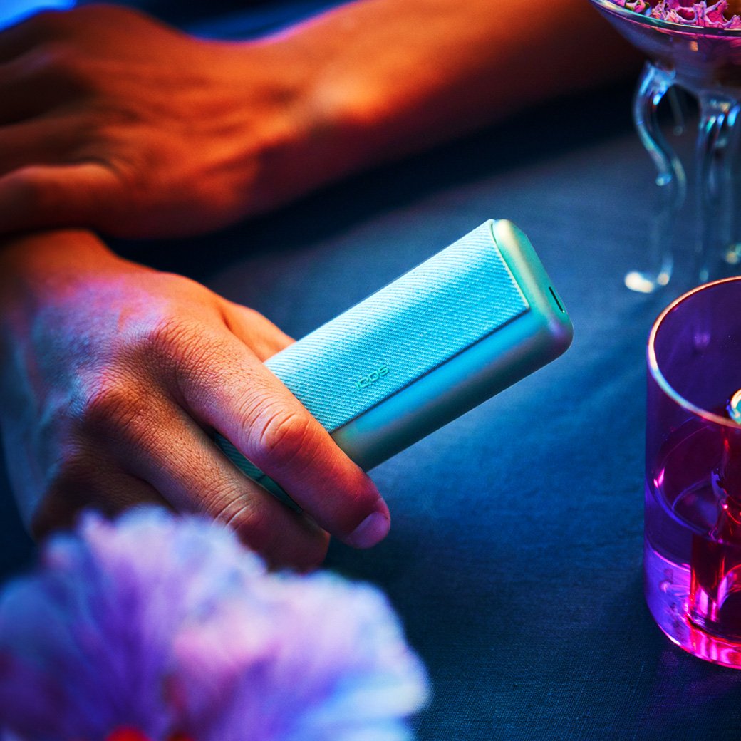 Two people holding mobile phones sit at a table; one also holds a jade blue IQOS ILUMA PRIME device.