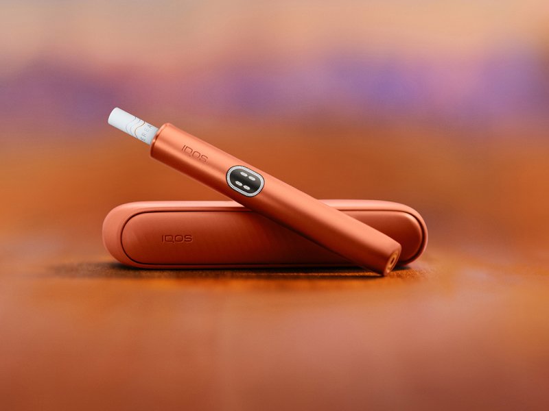 Zařízení IQOS ILUMA i v barvě Terracotta nad svou nabíječkou, s červeným pozadím