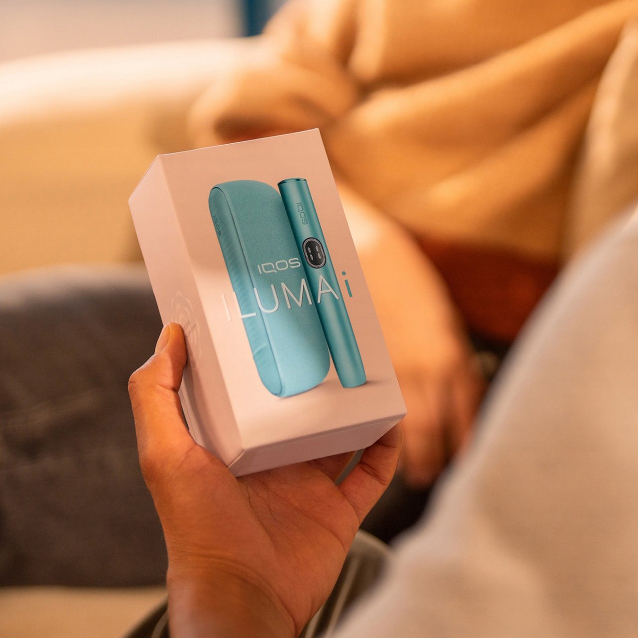 Eine Person hält eine Verpackung des IQOS ILUMA i Geräts in der Farbe Breeze Blue. Auf der Verpackung ist das Gerät abgebildet, bestehend aus einem schlanken, zylindrischen Element und einem Ladeetui. Der Text auf der Verpackung lautet ‚IQOS ILUMA i