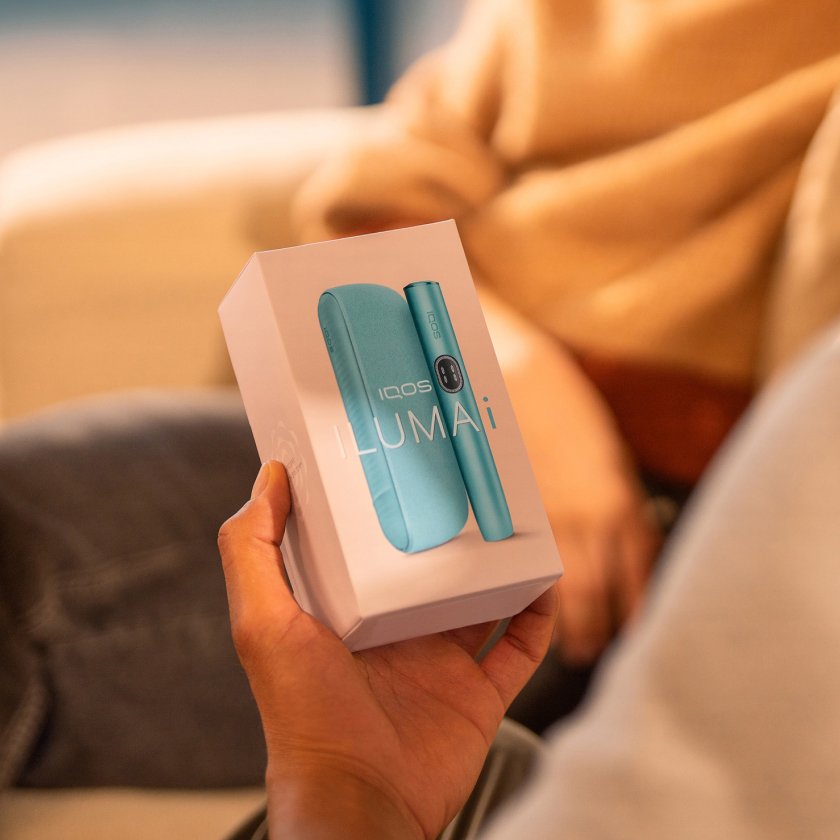 Eine Person hält eine Verpackung des IQOS ILUMA i Geräts in der Farbe Breeze Blue. Auf der Verpackung ist das Gerät abgebildet, bestehend aus einem schlanken, zylindrischen Element und einem Ladeetui. Der Text auf der Verpackung lautet ‚IQOS ILUMA i