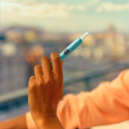 Hand holding an IQOS ILUMA i device, in front of a balcony