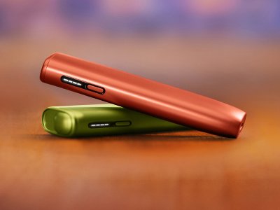 2 appareils IQOS ILUMA i ONE (appareil tout-en-un) couleurs terracotta et Vert Anis, posés l'un sur l'autre, sur un fond dégradé orange