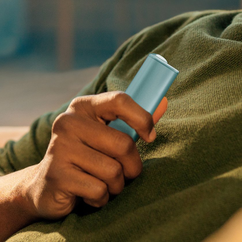 Eine Hand hält ein IQOS ILUMA i ONE Gerät in der Farbe Breeze Blue. Die Person trägt ein grünes Shirt und liegt oder lehnt sich zurück. Der Hintergrund ist unscharf, wodurch der Fokus auf dem Gerät liegt.