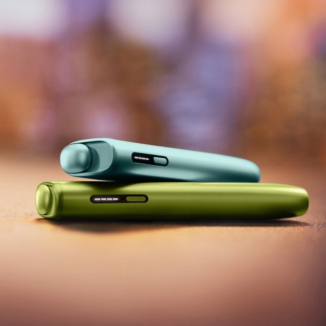 2 appareils IQOS ILUMA i ONE (appareil tout-en-un) couleurs Aigue Marine et Vert Anis, posés l'un sur l'autre, sur un fond dégradé orange