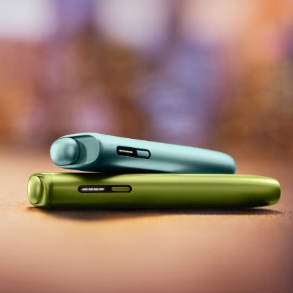 2 appareils IQOS ILUMA i ONE (appareil tout-en-un) couleurs Aigue Marine et Vert Anis, posés l'un sur l'autre, sur un fond dégradé orange