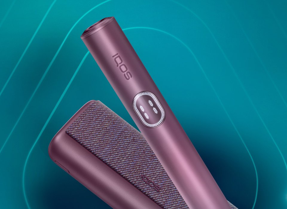 Main tenant un appareil IQOS ILUMA i PRIME turquoise; son chargeur posé sur une table.