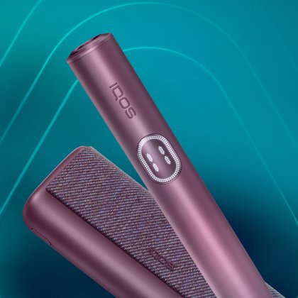 Main tenant un appareil IQOS ILUMA i PRIME turquoise; son chargeur posé sur une table.