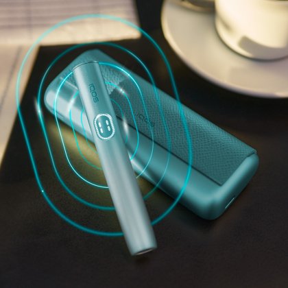 Hand holding a blue breezeIQOS ILUMA i PRIME device; its charger placed on a table