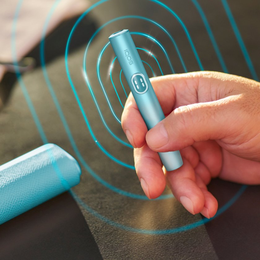 Hand holding a blue breezeIQOS ILUMA i PRIME device; its charger placed on a table