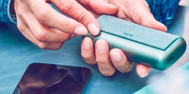 Hände halten ein IQOS ILUMA Prime über einem Smartphone an einem Tisch