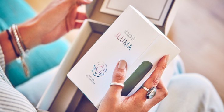 EIne Frau öffnet ein IQOS Paket und hält eine IQOS ILUMA Box in der Hand.