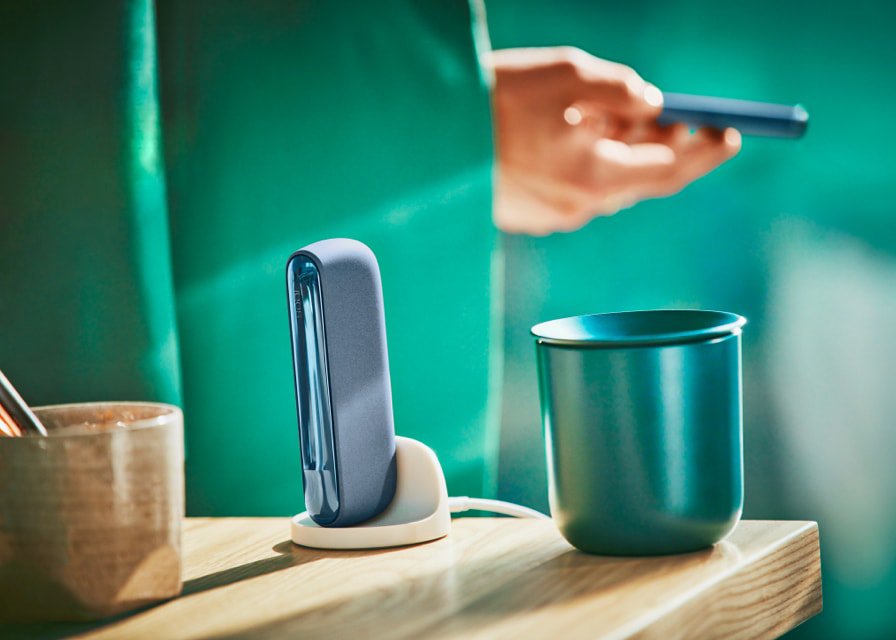 Man holds an IQOS ILUMA mid azure blue device, with a holder and cup on the table.