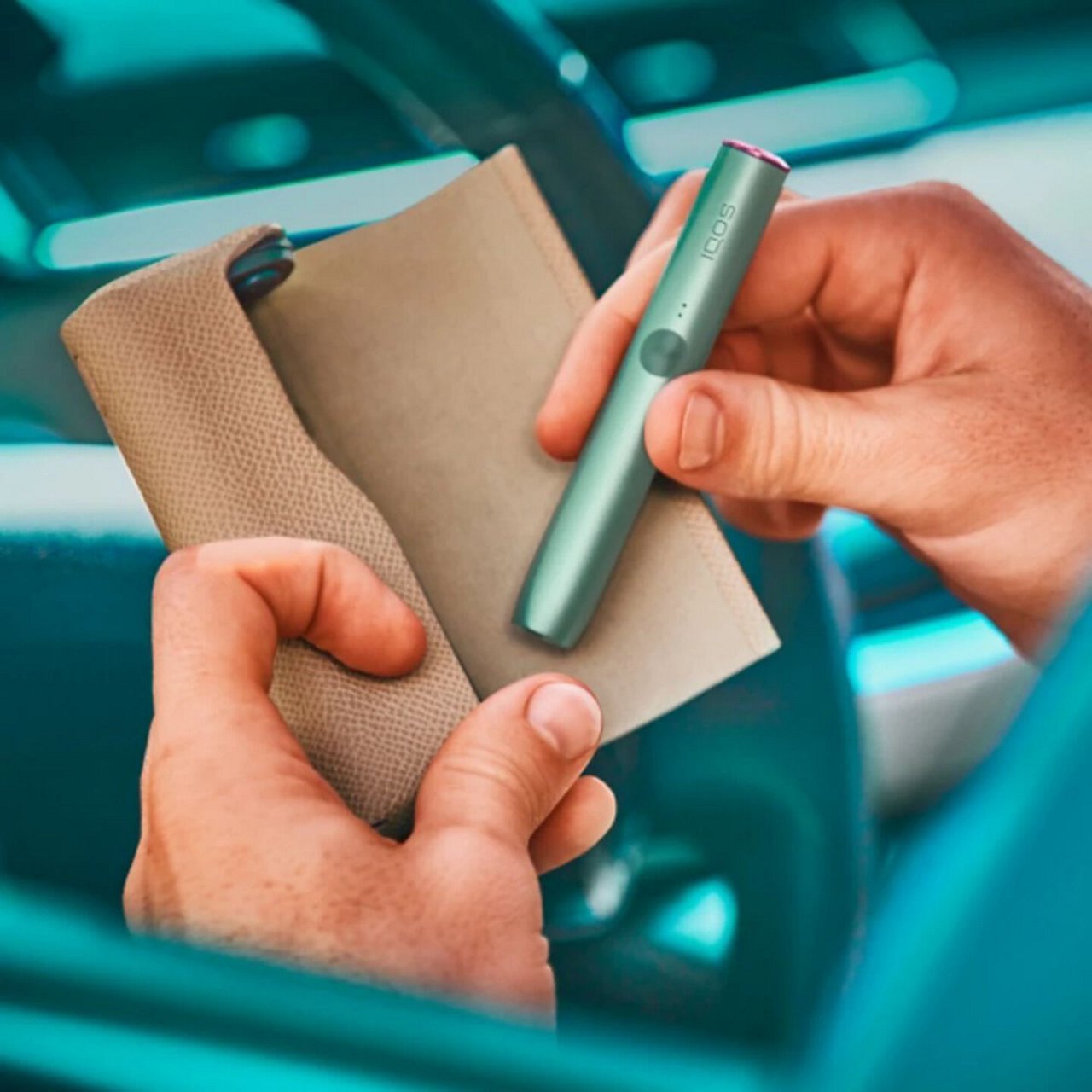 An IQOS device held by two hands in front of a car steering wheel