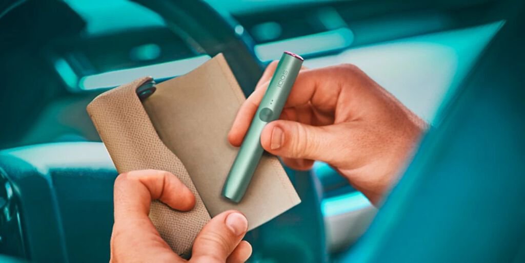 An IQOS device held by two hands in front of a car steering wheel