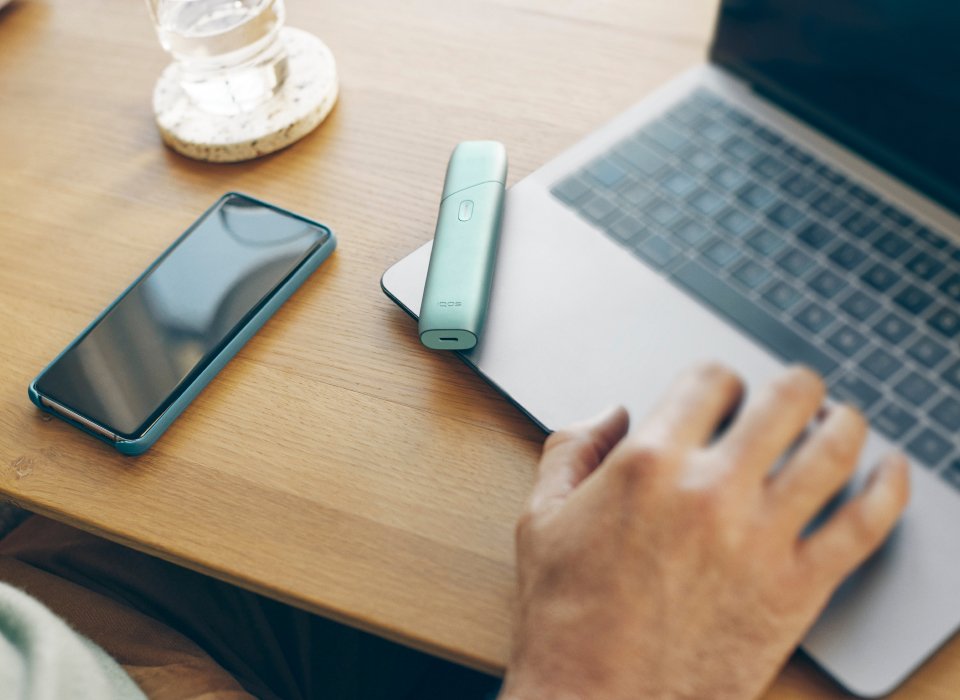  IQOS ILUMA ONE device placed on a laptop next to a mobile phone on a table.